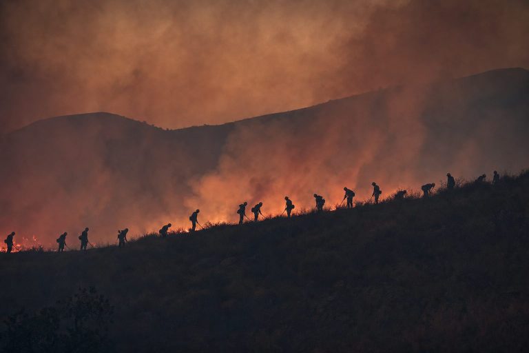A distant silhouetted hand crew working along a firebreak on a smoky ridgeline lit by ember glow